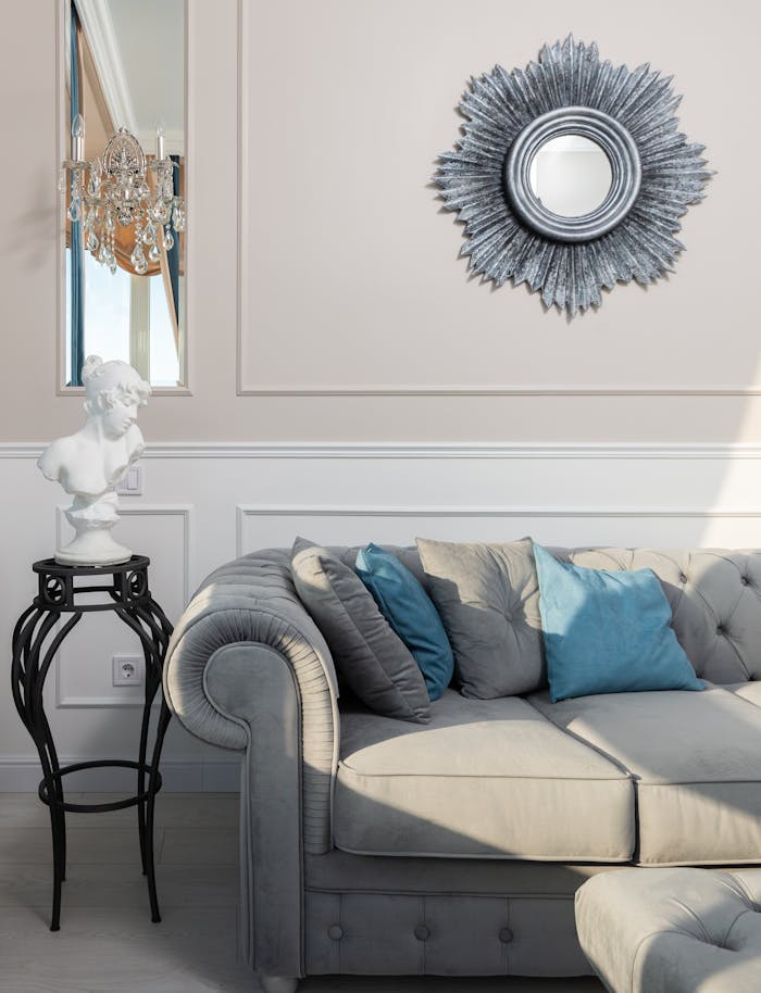 Elegant living room with a gray tufted sofa, blue accent pillows, ornate wall mirror, and classical bust statue, creating a sophisticated home decor style